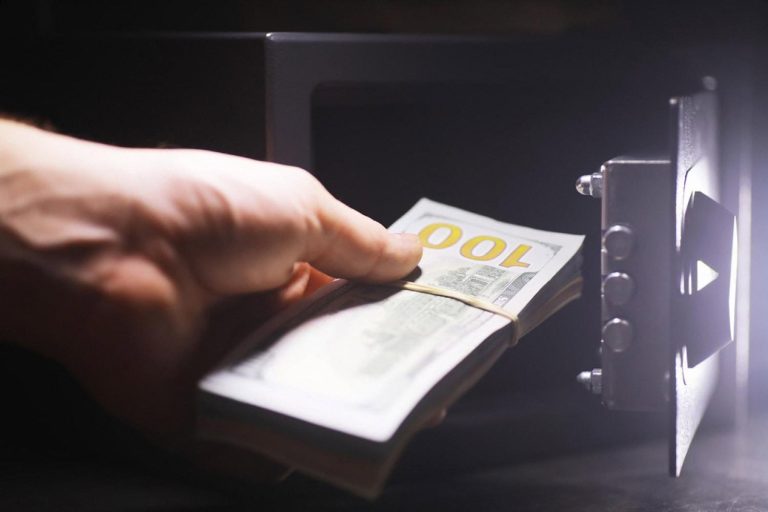 Hand placing a stack of US dollar bills into a small home safe with an electronic lock