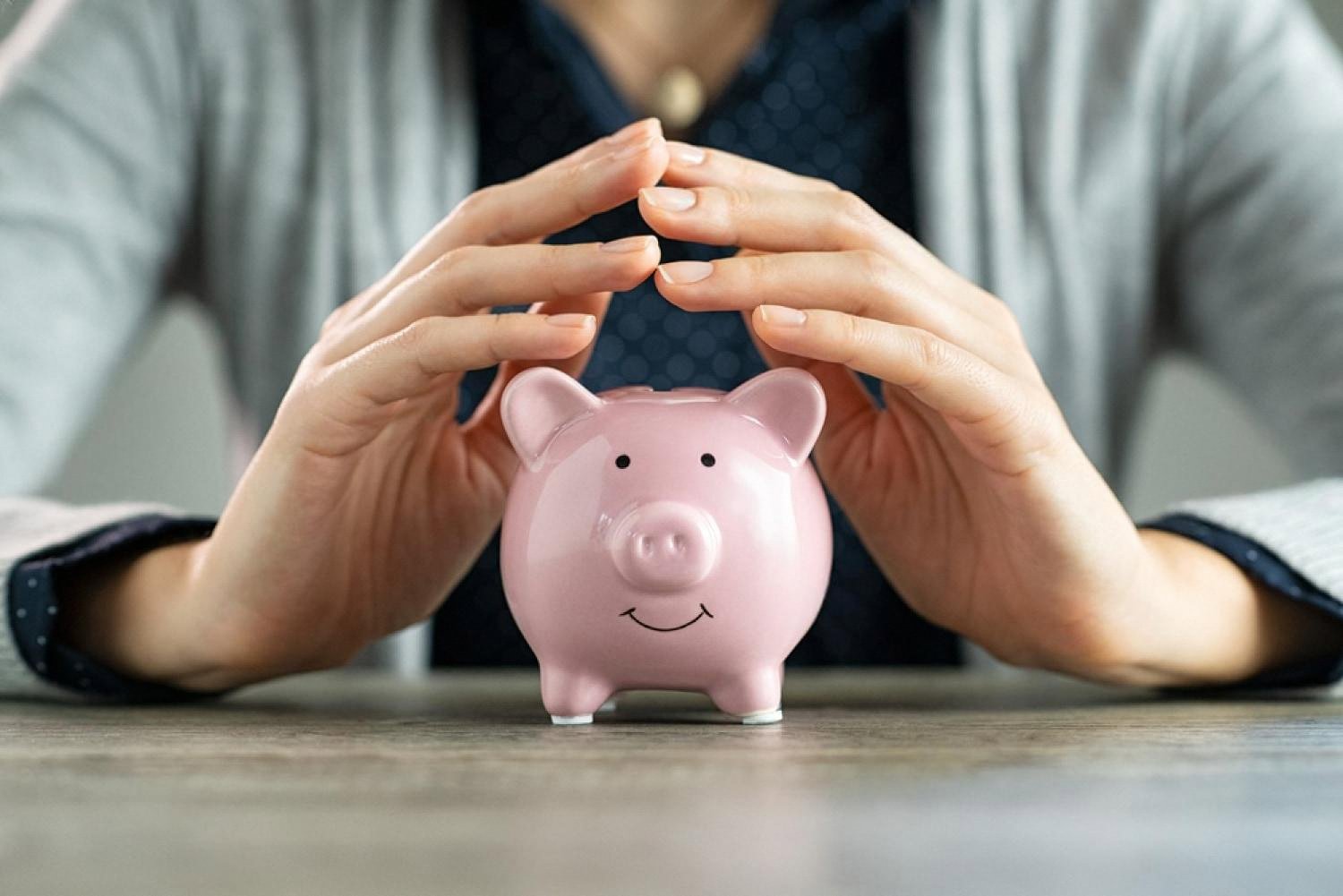 Two hands shielding a pink piggy bank on a wooden table, symbolising home savings protection